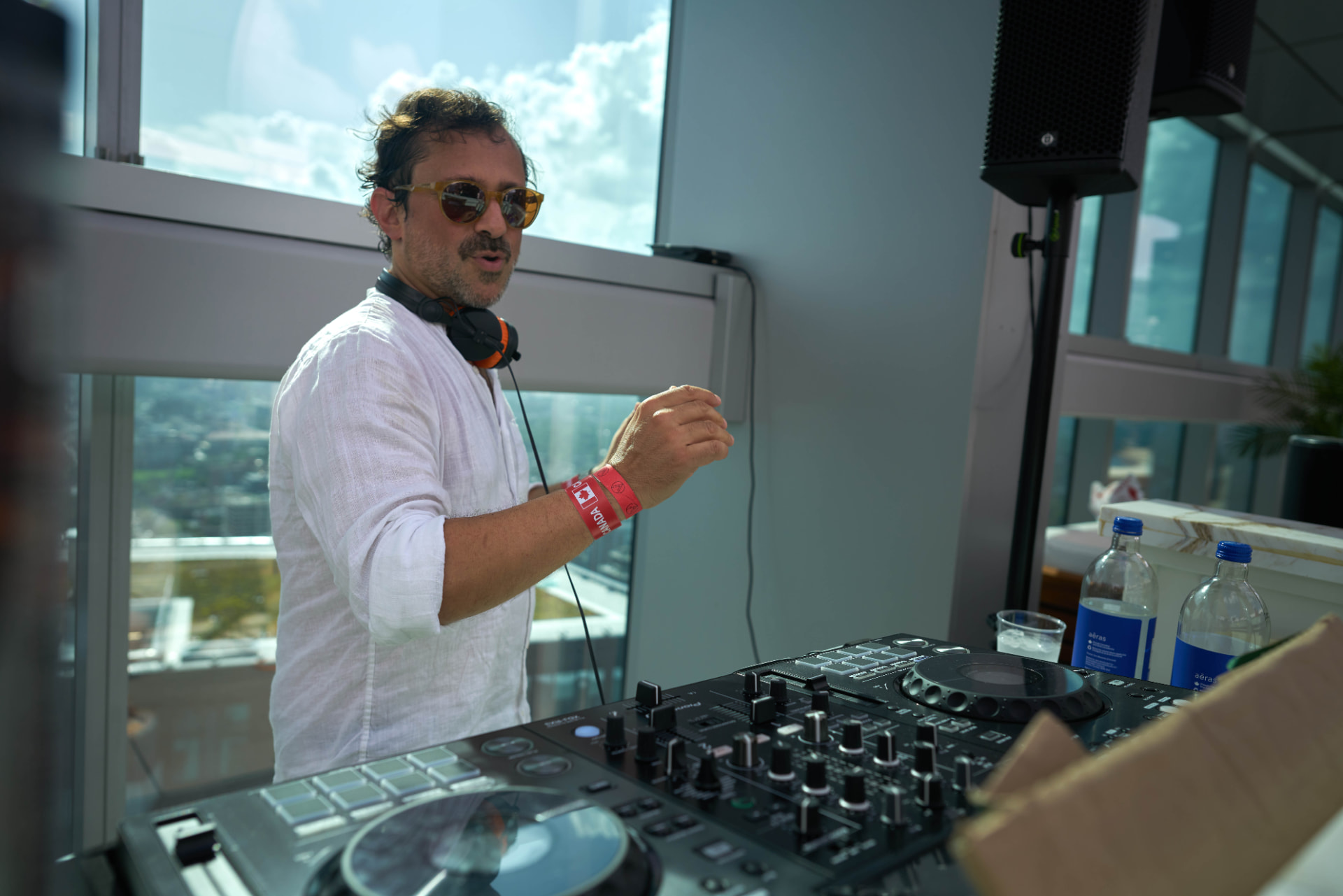 DJ in a white linen shirt cueing a track beside the speaker stack with the skyline behind