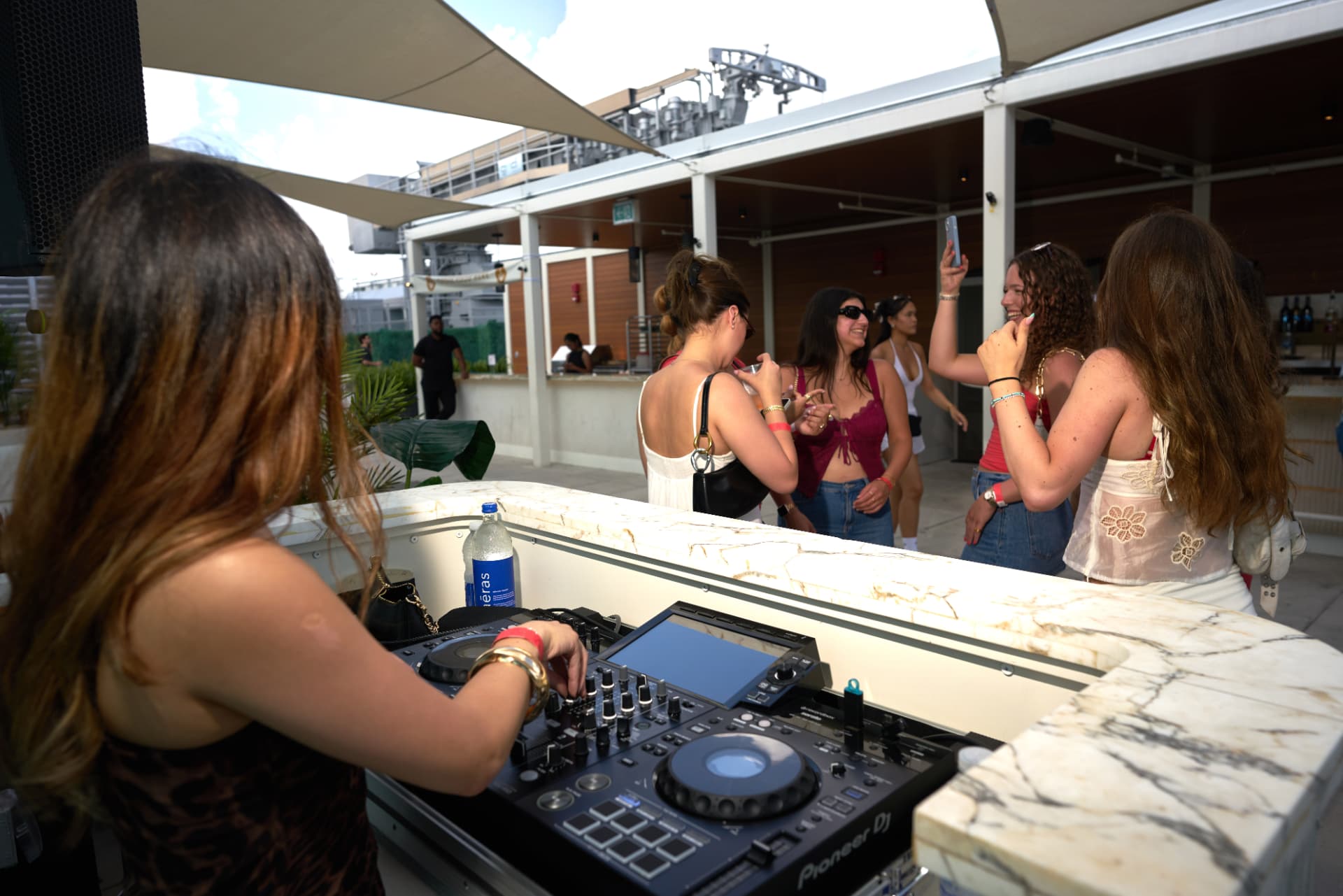 DJ behind the marble bar as friends dance and film in the afternoon sun